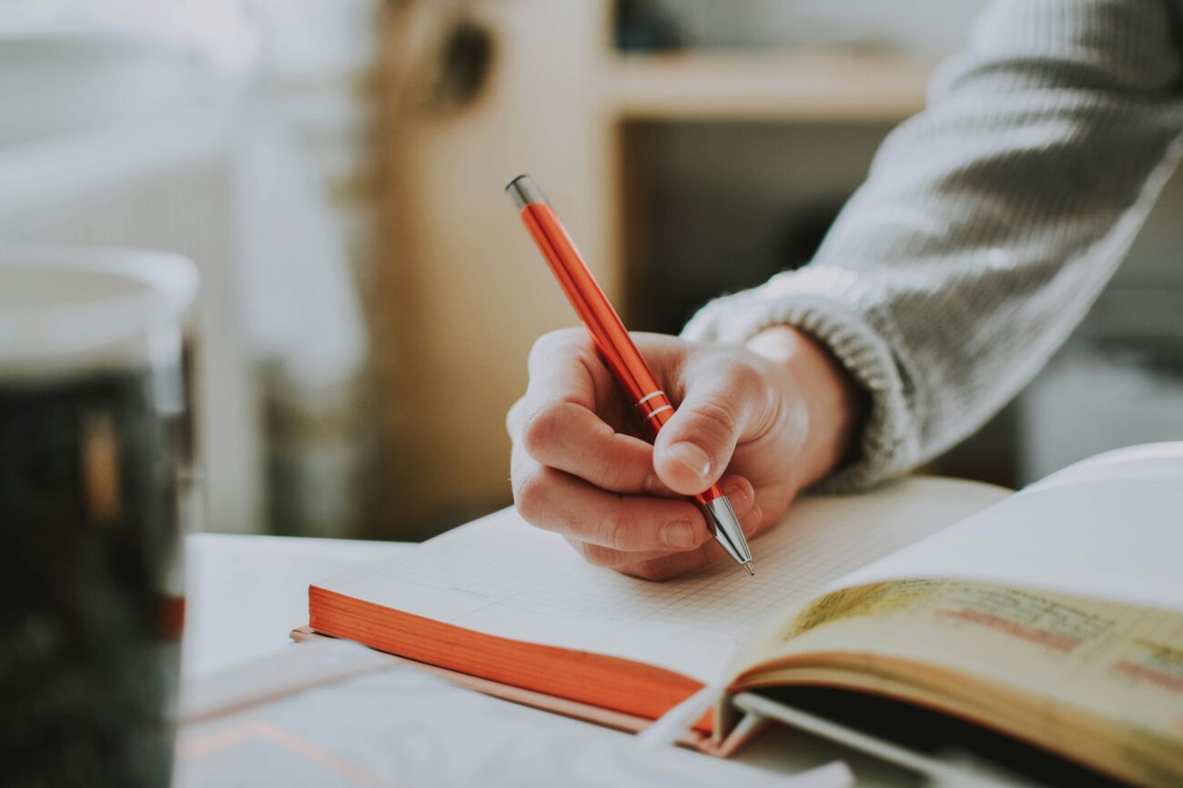 A hand writing with a red pen in an open notebook on a table by a window