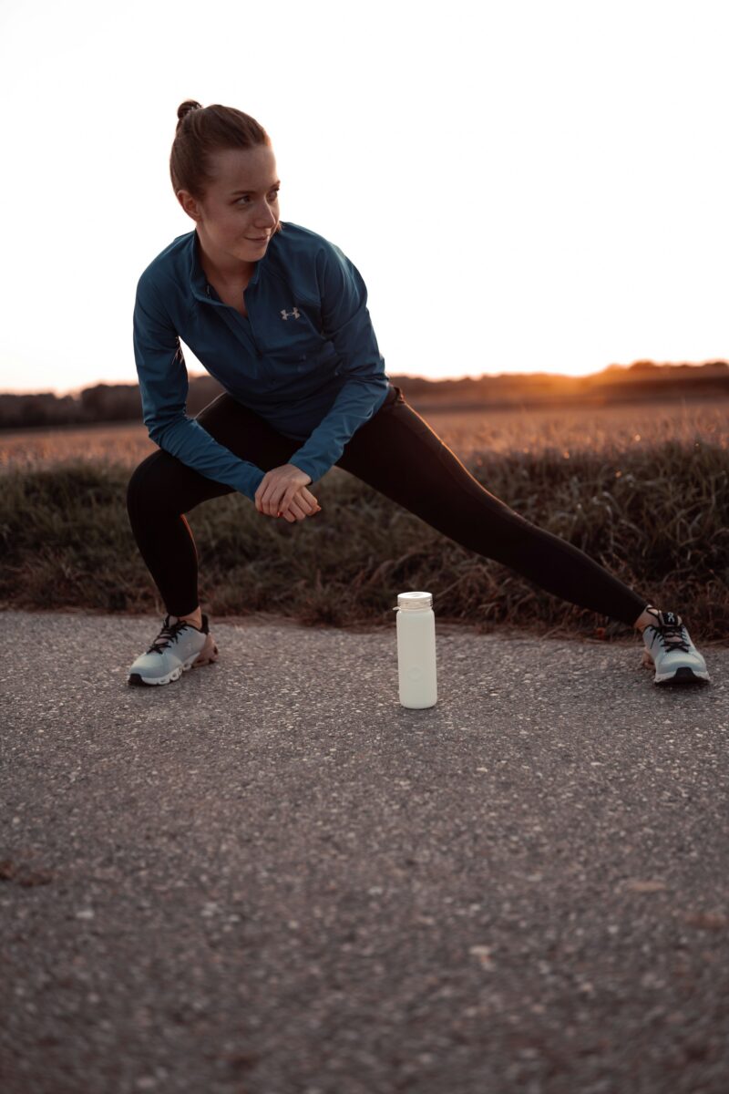 A woman in running clothes doing a side lunge stretch on a path at sunset, a water bottle on the ground beside her