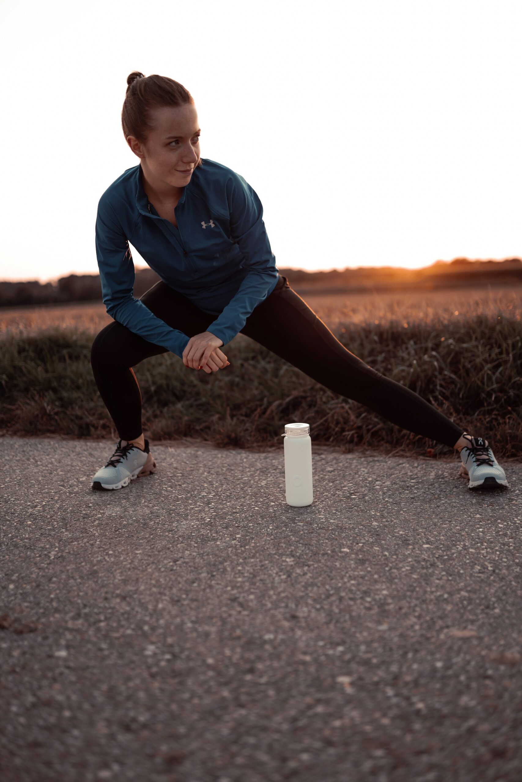 A woman in running clothes doing a side lunge stretch on a path at sunset, a water bottle on the ground beside her