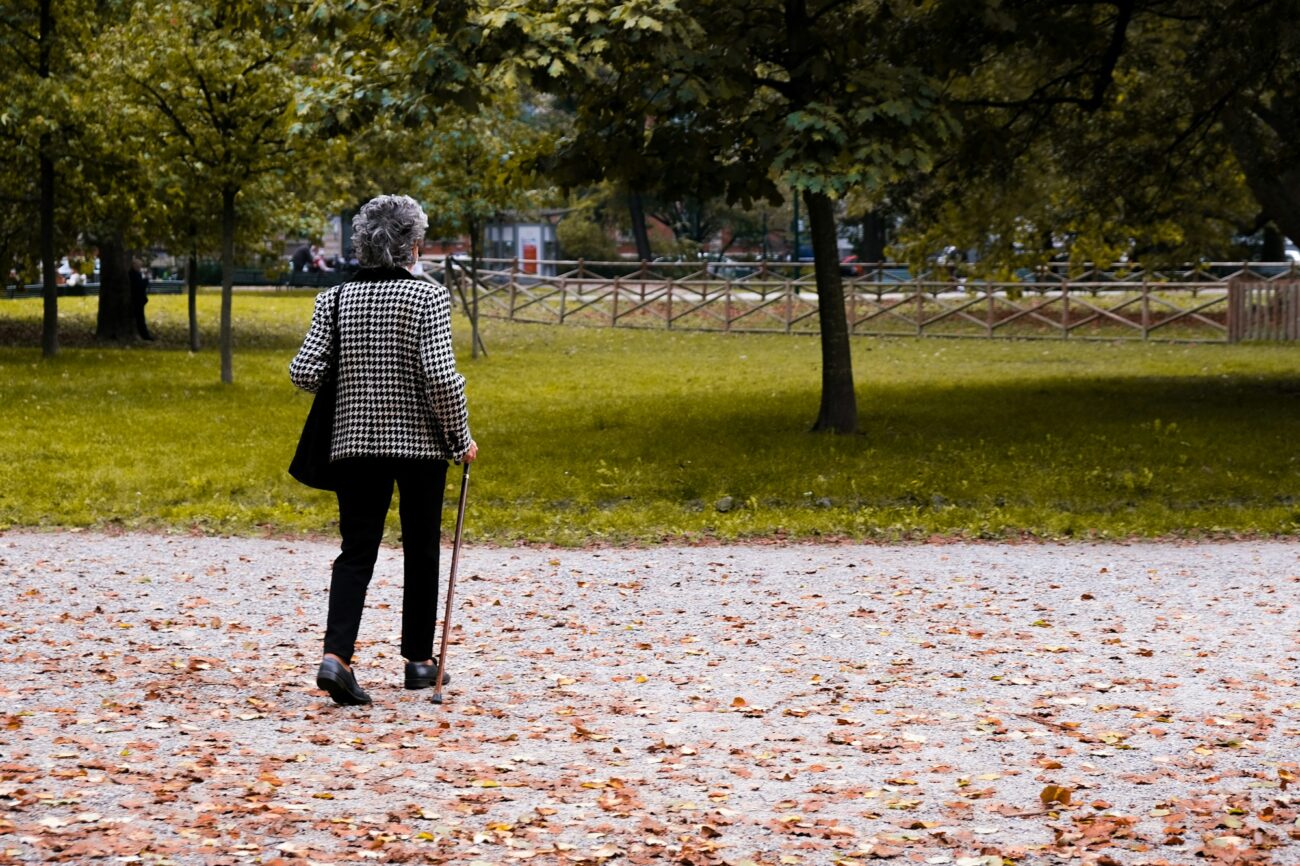 An older woman with a walking cane taking a walk along a leaf-covered park path in autumn
