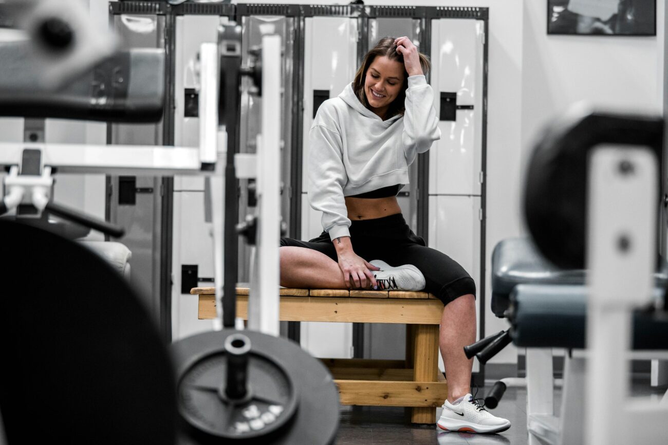 A woman in a hoodie sitting on a wooden gym bench between workout machines, resting after a session