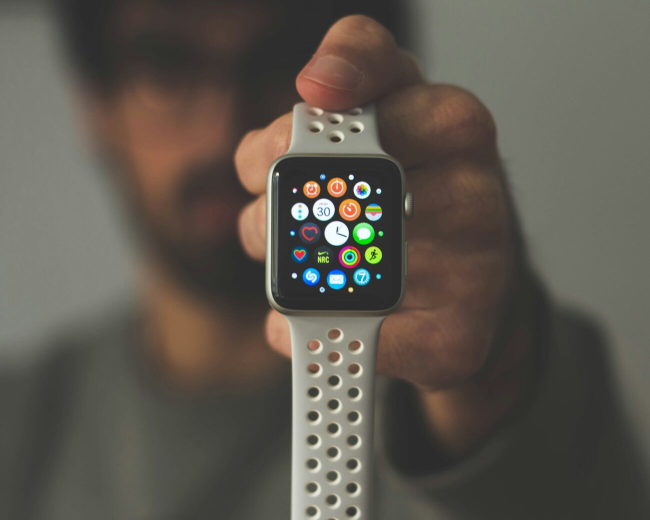 A man holding a smartwatch toward the camera, its screen full of app icons, his face blurred behind