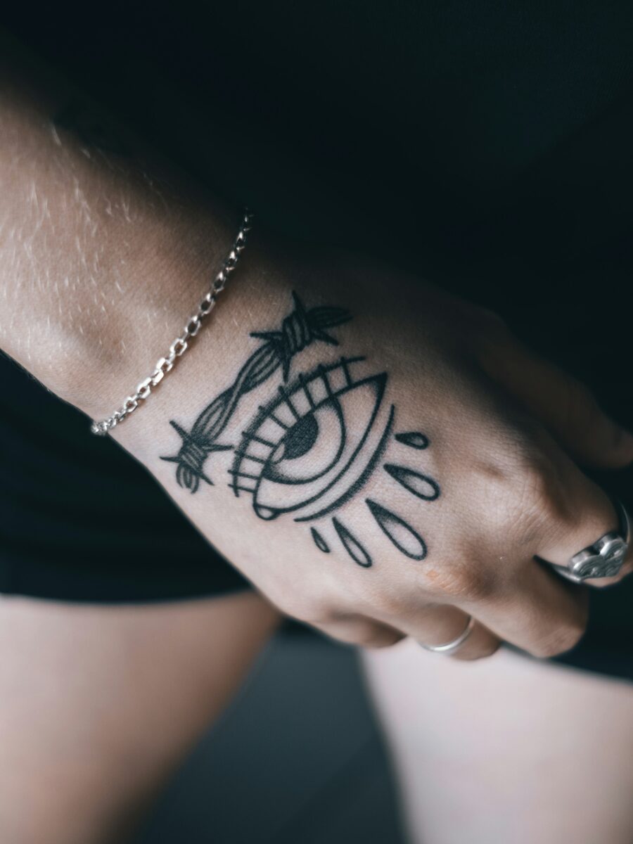 A black ink tattoo of an eye across the back of a hand and wrist, a thin chain bracelet above it