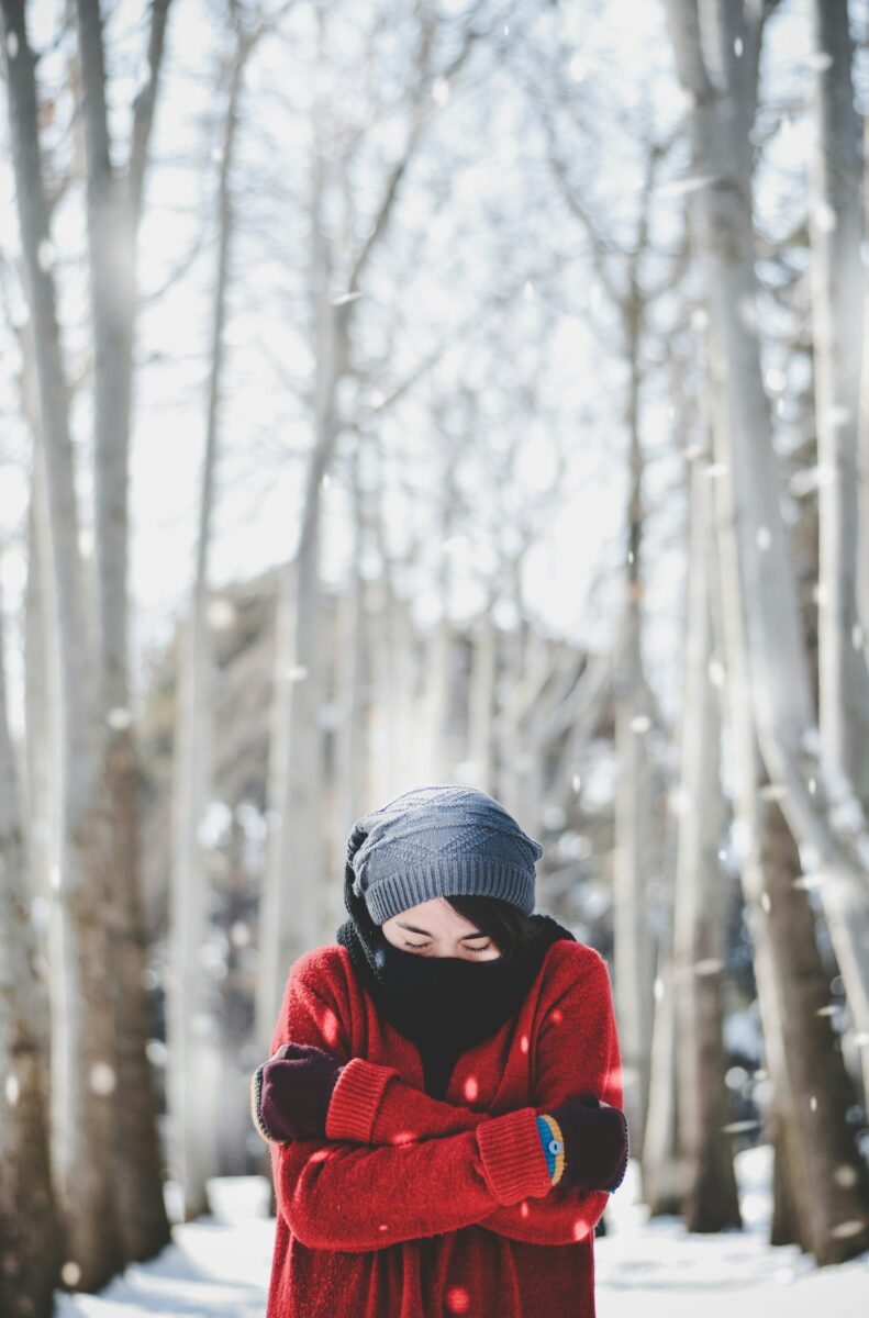 A woman in a red coat, scarf, knit cap and mittens hugging herself against the cold in a snowy row of trees
