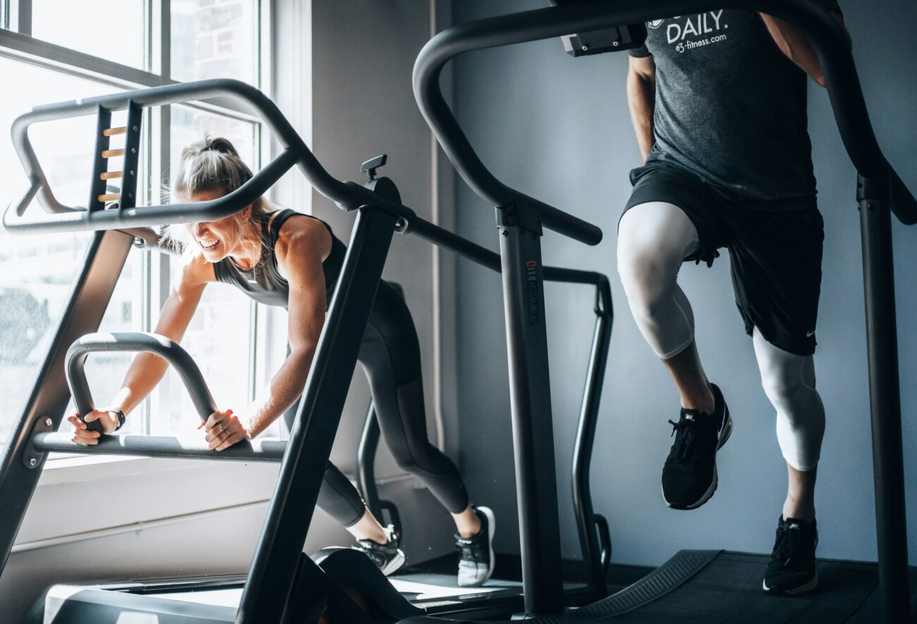 Two people working out side by side on treadmills in a bright gym by a window
